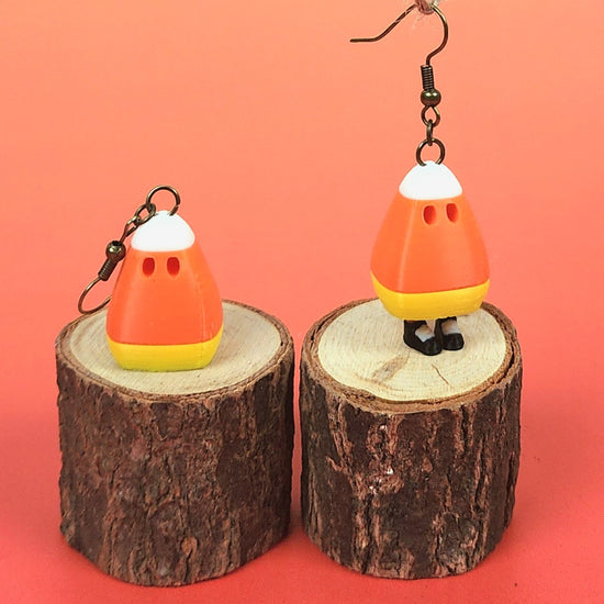 A candy corn attached to an earring hook is shown dangling from a string. It is begins turned away from the camera and resting on a small piece of wood. As the string lifts, the candy corn lifts up to reveal little legs and feet that dangle out from beneath it. The candy corn spins around a bit, showing its little eyes and dancing to a happy tune. The candy corn then jumps out of the video frame.